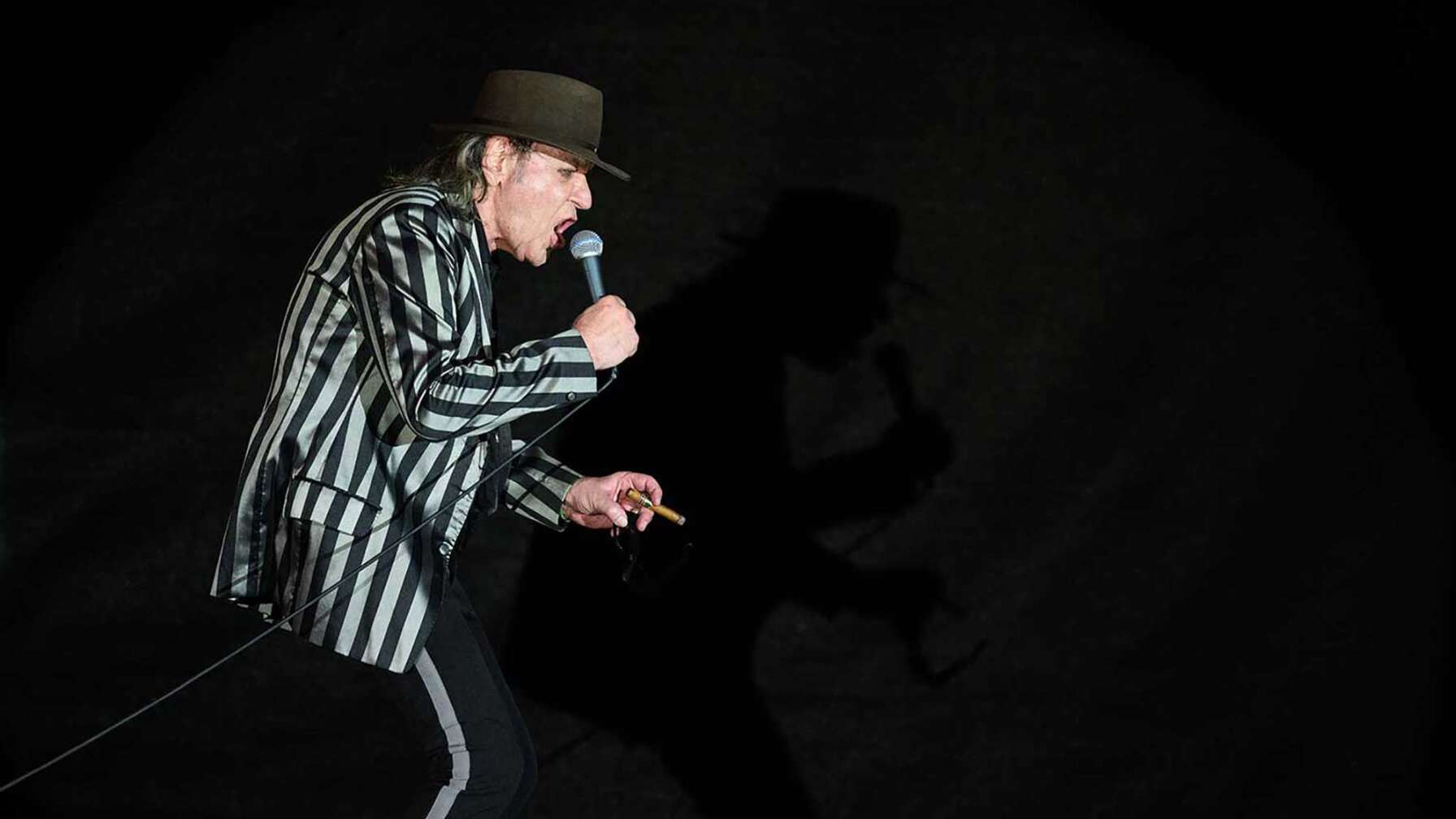 Sänger Udo Lindenberg performt mit Mikrofon in der Hand und Hut auf dem Kopf auf der Bühne.