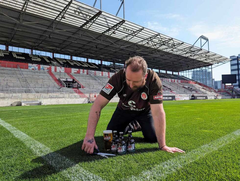 ASTRA Chefbrauer Merlin Monagan kniet auf der Rasenfläche im Millerntorstadion und schneidet Gras ab