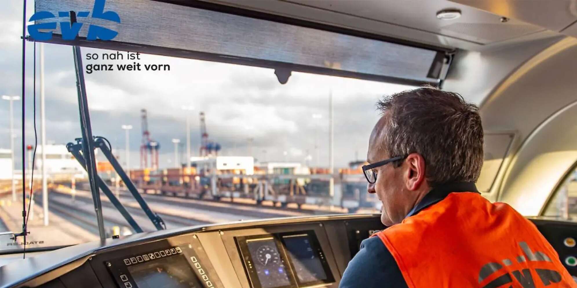 Ein Mann sitzt im Cockpit einer der Züge der EVB und blickt auf die Schienen, im Hintergrund der Hamburger Hafen