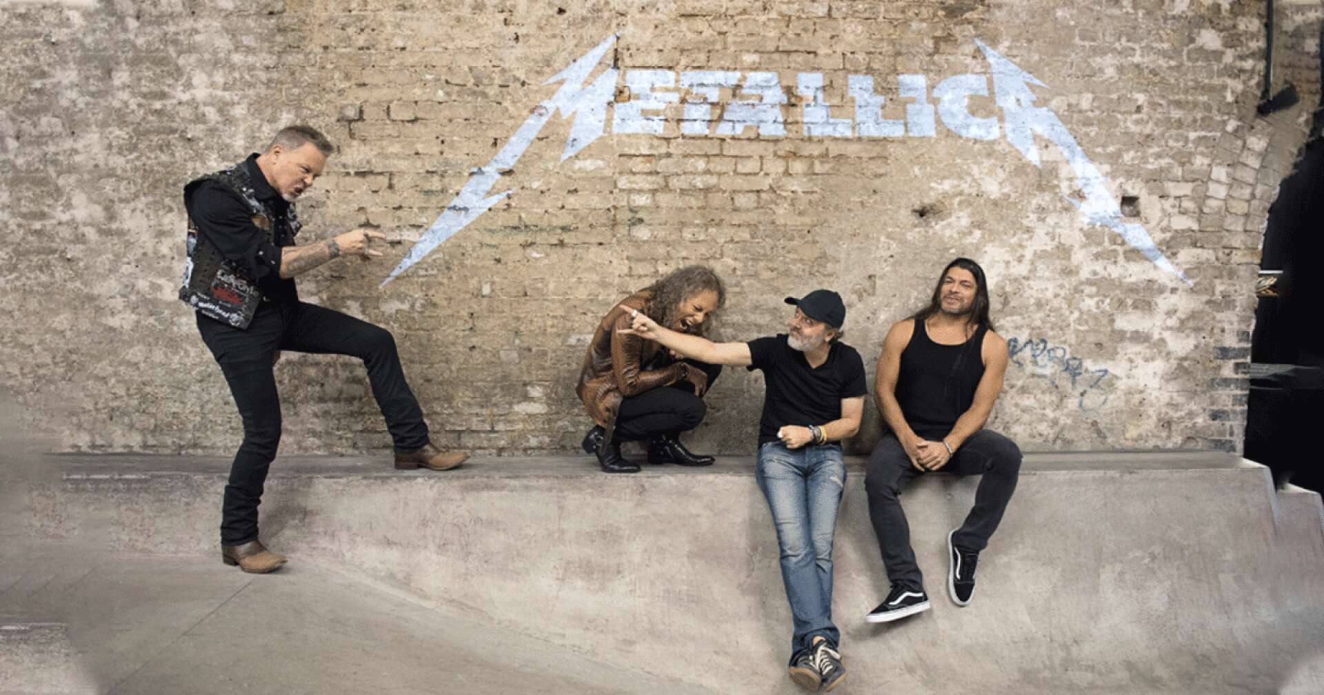 Metallica posieren vor einer Backstein mit ihrem Logo